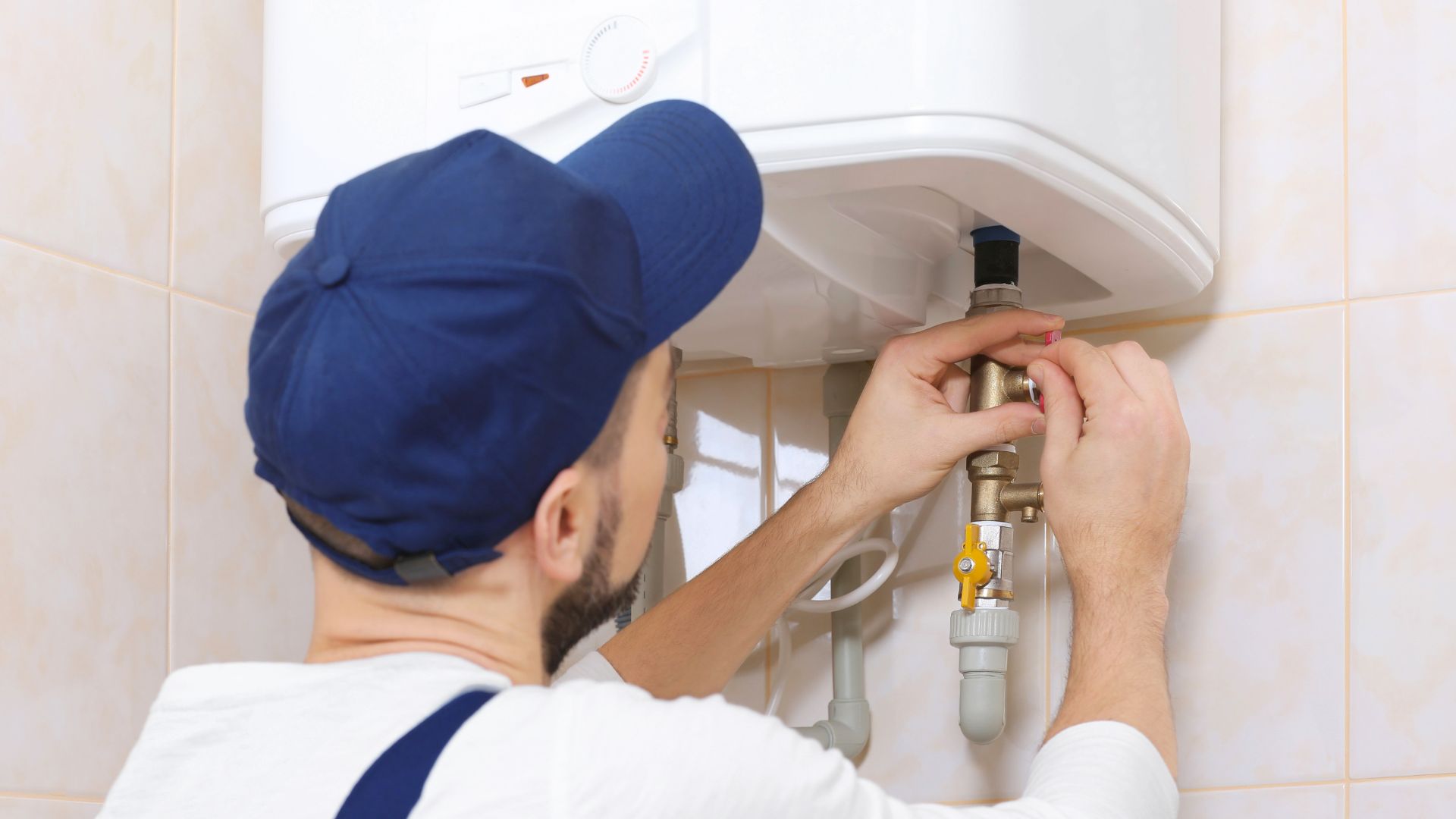 Plumber in blue cap fixing water heater pipes in tiled bathroom