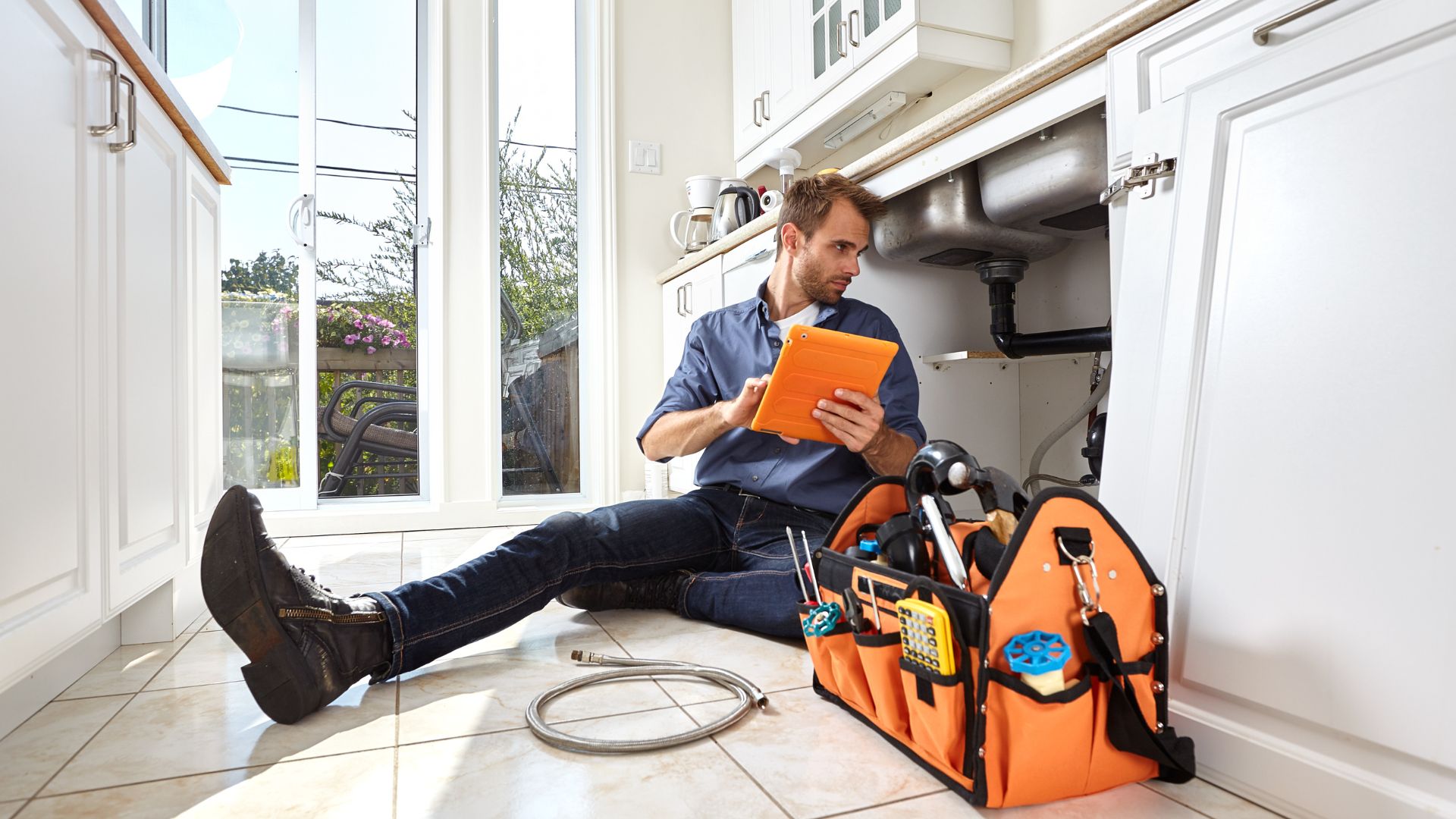 Plumber checking tablet while sitting on kitchen floor with tools nearby