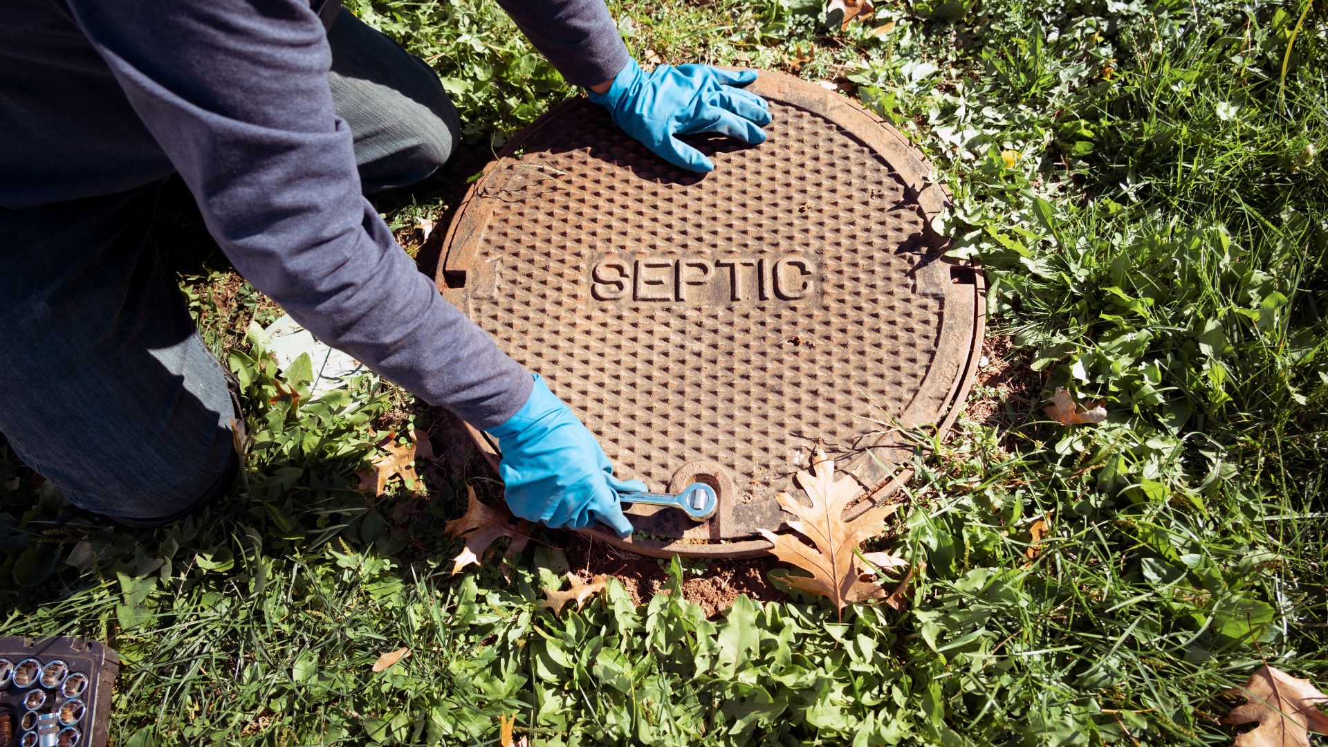 Gloved hands opening a rusty septic tank lid surrounded by grass and leaves