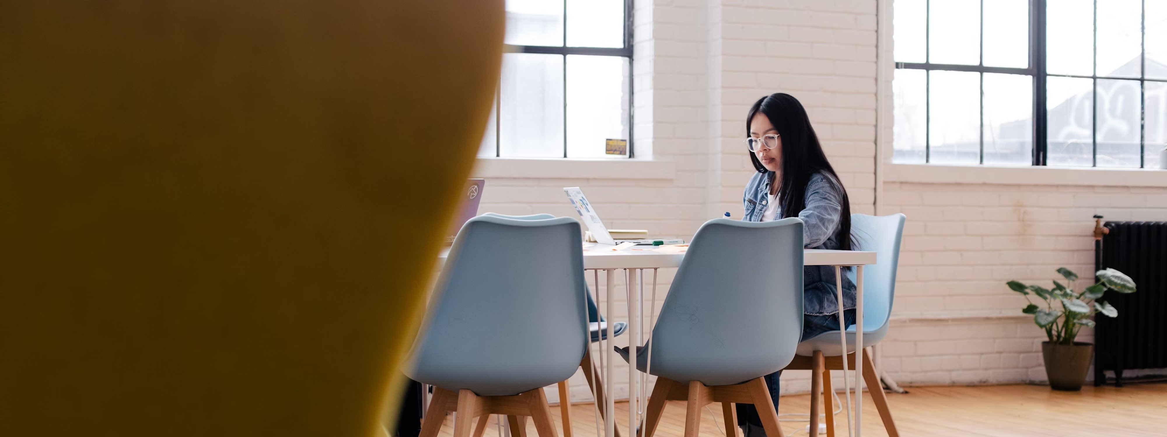 Woman working on laptop in bright, minimalist workspace with blue chairs