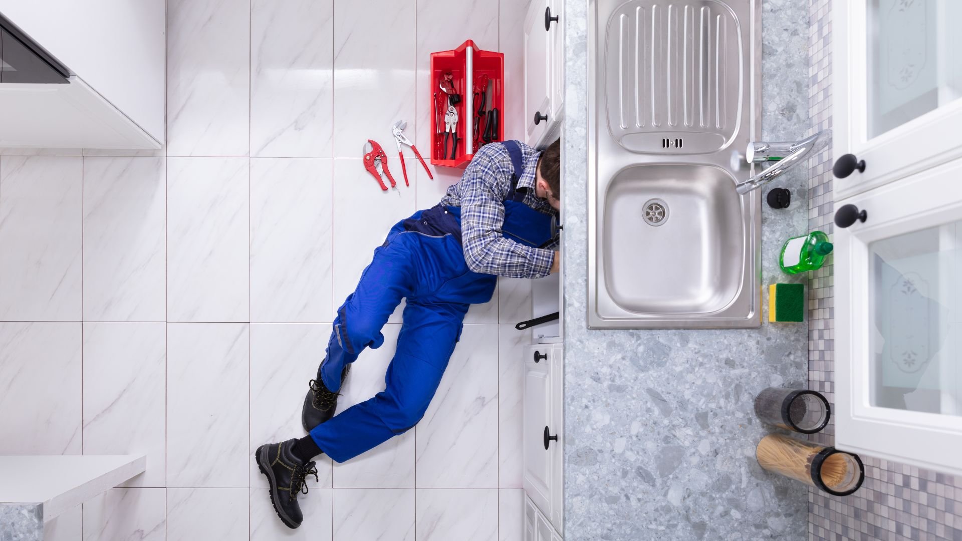 Plumber fixing sink under kitchen counter with red toolbox nearby